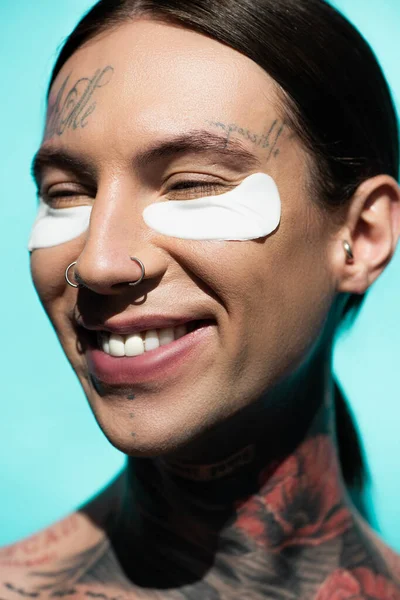 Primer plano del joven tatuado con parches en los ojos sonriendo aislado en turquesa - foto de stock