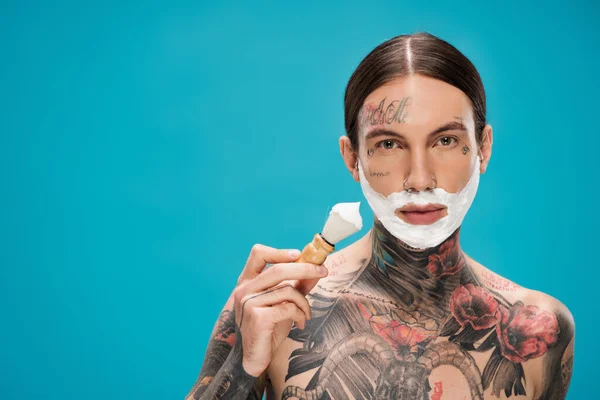 Hombre tatuado y sin camisa con espuma de afeitar en la cara con cepillo vintage aislado en azul - foto de stock