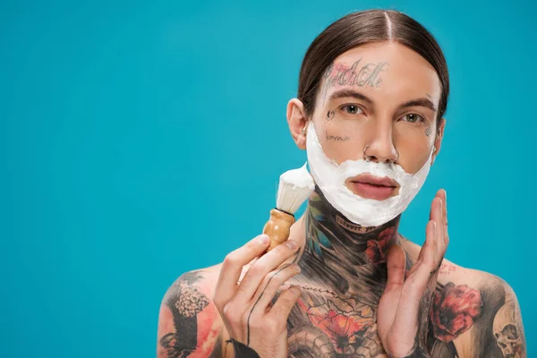 Homem tatuado com espuma de barbear no rosto segurando escova vintage isolado em azul — Fotografia de Stock