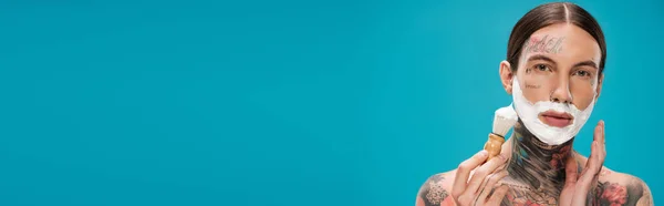 Young man with tattoos and shaving foam on face holding vintage brush isolated on blue, banner — Stock Photo