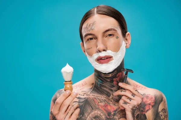 Tattooed young man with shaving foam on face holding vintage brush and safety razor isolated on blue — Stock Photo