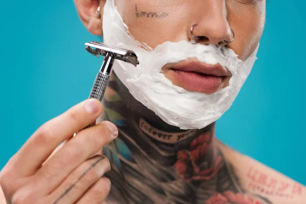 Cropped view of tattooed young man with shaving foam on face holding safety razor isolated on blue — Stock Photo