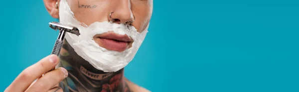 Vista ritagliata del giovane tatuato con schiuma da barba sul viso tenuta rasoio di sicurezza isolato su blu, banner — Foto stock