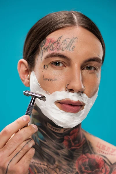 Tattooed young man with shaving foam on face holding safety razor isolated on blue — Stock Photo