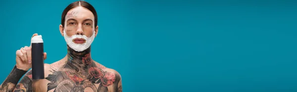 Tattooed young man with shaving foam on face holding can isolated on blue, banner — Stock Photo