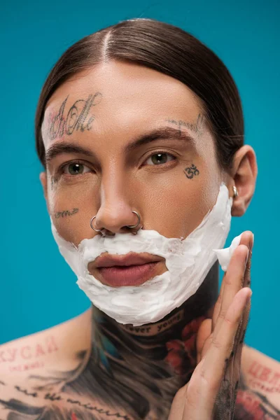 Tatuato giovane uomo applicando schiuma da barba bianca sul viso isolato su blu — Foto stock