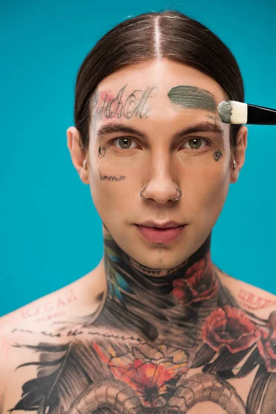 Jeune homme tatoué appliquant masque en argile avec pinceau cosmétique isolé sur bleu — Photo de stock