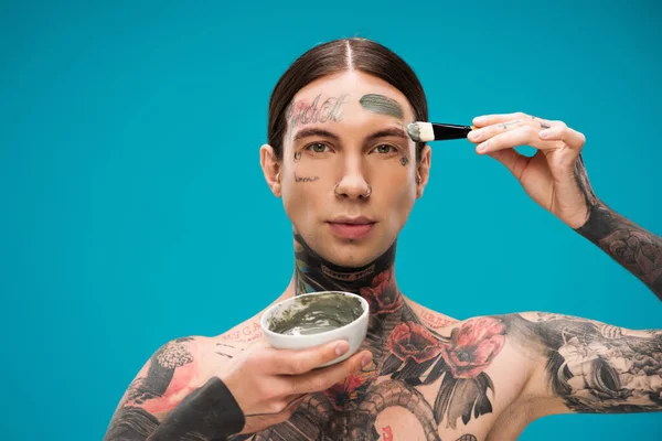 Tattooed young man applying clay mask with cosmetic brush and holding bowl with product isolated on blue — Stock Photo