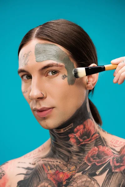 Pierced young man with tattoos applying clay mask with cosmetic brush isolated on blue — Stock Photo