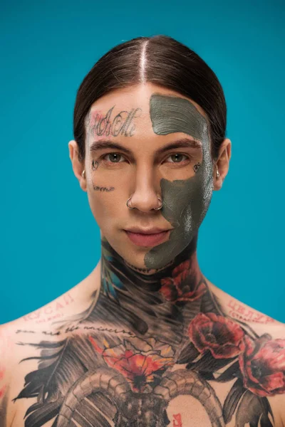 Percé jeune homme avec tatouages et masque d'argile sur le visage en regardant la caméra isolée sur bleu — Photo de stock