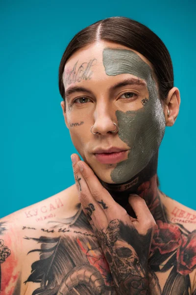 Jeune homme avec tatouages et masque d'argile sur le visage en regardant la caméra isolée sur bleu — Photo de stock