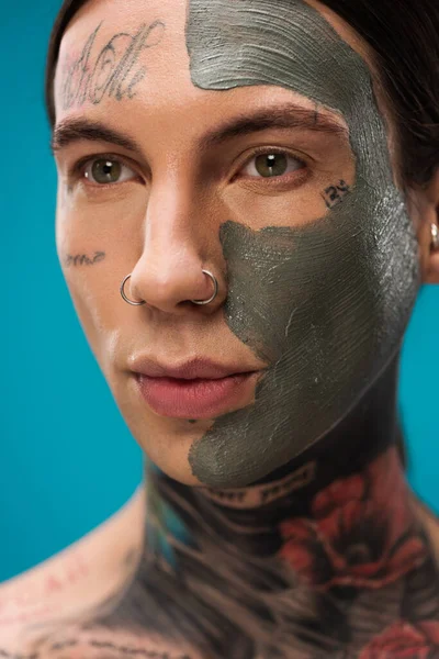 Gros plan de jeune homme percé de tatouages et masque d'argile sur le visage isolé sur bleu — Photo de stock