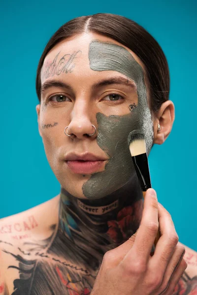 Jovem perfurado com tatuagens aplicando máscara de barro no rosto com escova cosmética isolada em azul — Fotografia de Stock