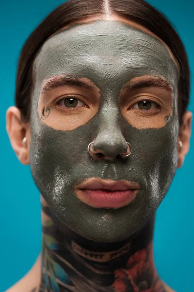 Close up of pierced young man with tattoos and clay mask on face isolated on blue — Stock Photo