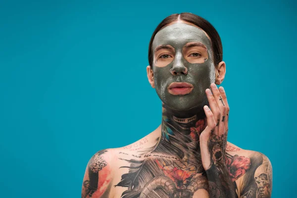 Без сорочки молодий чоловік з татуюваннями торкається обличчя з глиняною маскою ізольовано на синьому — стокове фото