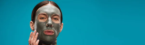 Молодой человек с татуировками трогательное лицо с глиняной маской изолированы на синий, баннер — стоковое фото