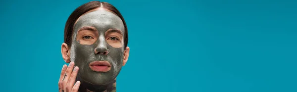 Joven con tatuajes tocando la cara con máscara de arcilla aislado en azul, pancarta - foto de stock