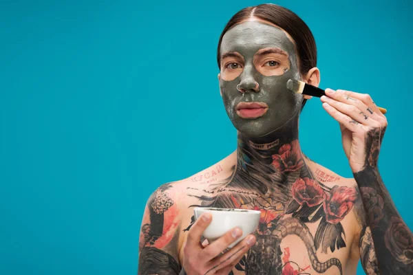 Shirtless young man with tattoos applying clay mask with cosmetic brush and holding bowl isolated on blue — Stock Photo