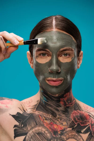 Hombre joven sin camisa con tatuajes aplicando máscara de arcilla con cepillo cosmético aislado en azul - foto de stock