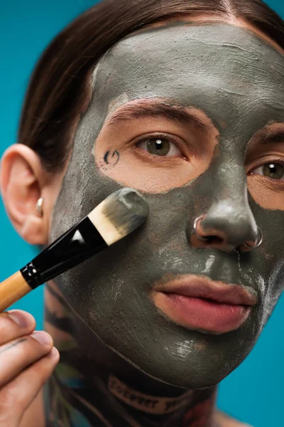 Primo piano del giovane trafitto con tatuaggi applicare maschera di argilla con pennello cosmetico isolato su blu — Foto stock