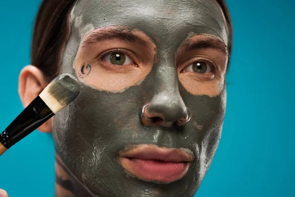 Nahaufnahme eines gepiercten jungen Mannes, der Tonmaske mit Kosmetikpinsel auf blauem Grund aufträgt — Stockfoto