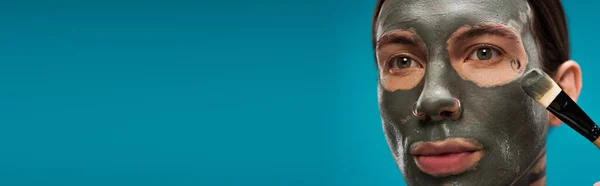 Close-up de jovem perfurado aplicando máscara de barro com escova cosmética isolada em azul, banner — Fotografia de Stock