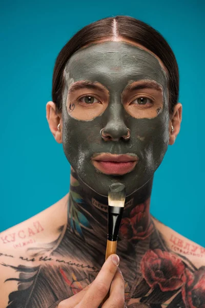 Пронзительный человек с татуировками наносит глиняную маску с косметической щеткой, изолированной на голубом — стоковое фото