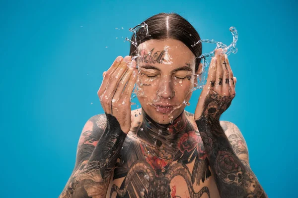 Spruzzata d'acqua vicino giovane tatuato uomo lavaggio viso isolato su blu — Foto stock