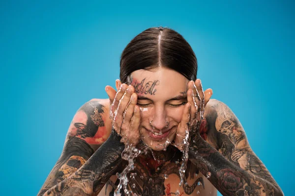 Spruzzata d'acqua vicino all'uomo felice e tatuato che lava il viso isolato sul blu — Foto stock