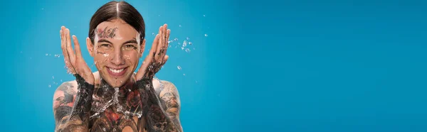 Splash of water near cheerful man with tattoos washing face isolated on blue, banner — Stock Photo