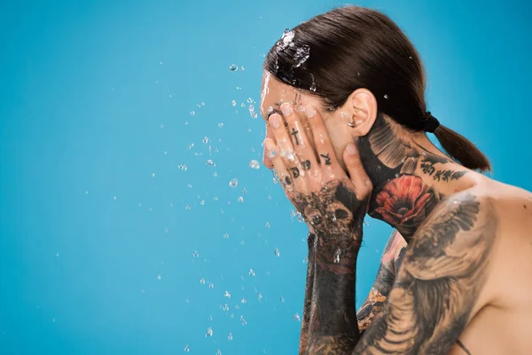 Éclaboussure d'eau près de l'homme avec des tatouages lavage visage isolé sur bleu — Photo de stock