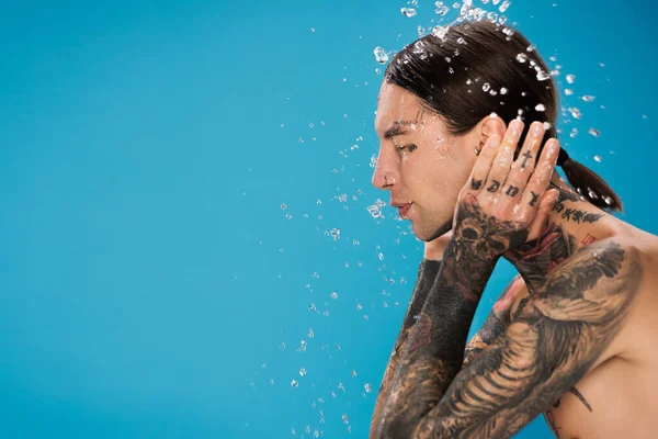 Salpicadura de agua cerca de un joven con tatuajes y ojos cerrados lavando la cara aislada en azul - foto de stock