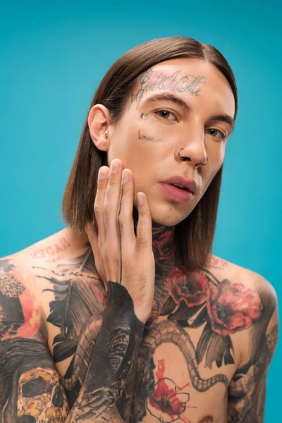 Без сорочки молодий чоловік з татуюваннями дивиться на камеру, торкаючись гладкої шкіри, ізольованої на синьому — стокове фото