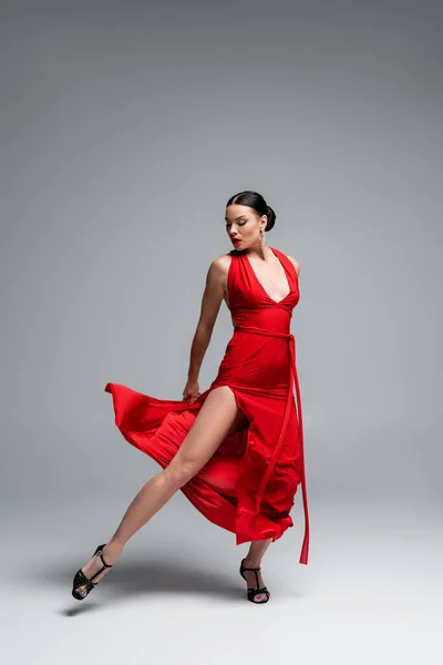 Full length of brunette dancer in red dress looking at leg on grey background — Stock Photo