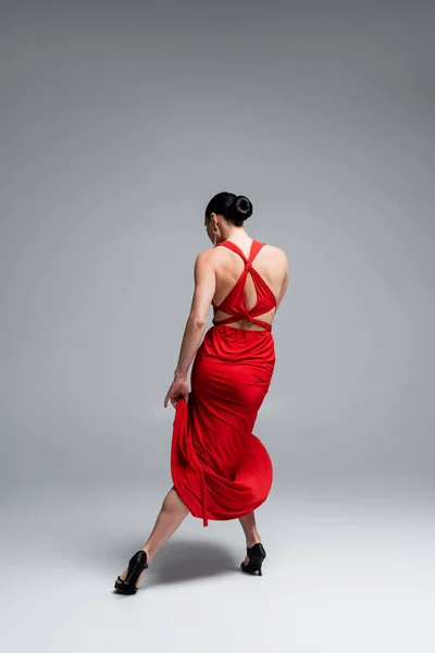 Vista trasera de mujer morena en vestido rojo y tacones bailando tango sobre fondo gris - foto de stock