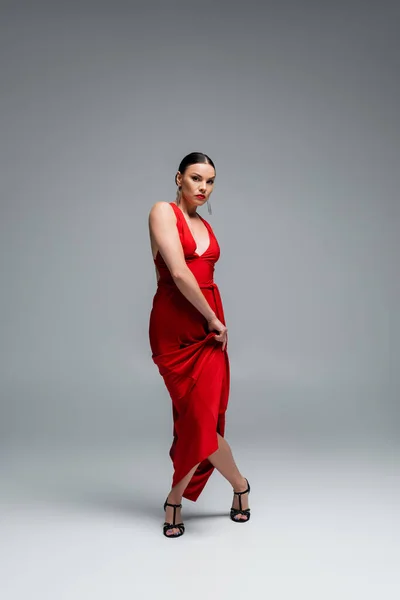 Стильный бальный танцор в красном платье и на каблуках смотрит в камеру на сером фоне — стоковое фото
