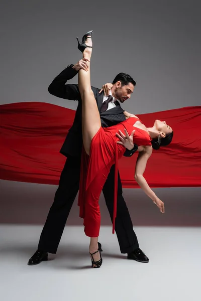 Vue latérale du danseur élégant touchant la jambe du partenaire tout en effectuant le tango sur fond gris avec tissu rouge — Photo de stock