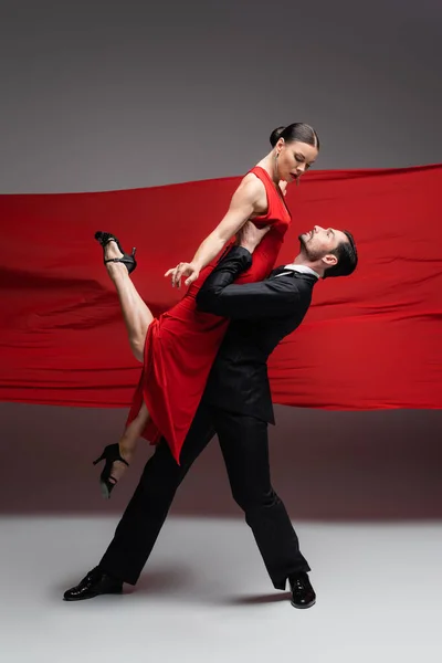 Danseuse en costume soulevant partenaire élégant tout en effectuant tango sur fond gris avec tissu rouge — Photo de stock