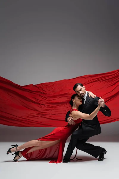 Elegant couple holding hands while performing tango on grey background with red fabric — Stock Photo