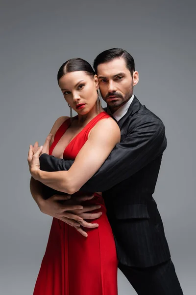 Uomo in abito che abbraccia donna elegante in abito rosso mentre balla tango isolato sul grigio — Foto stock