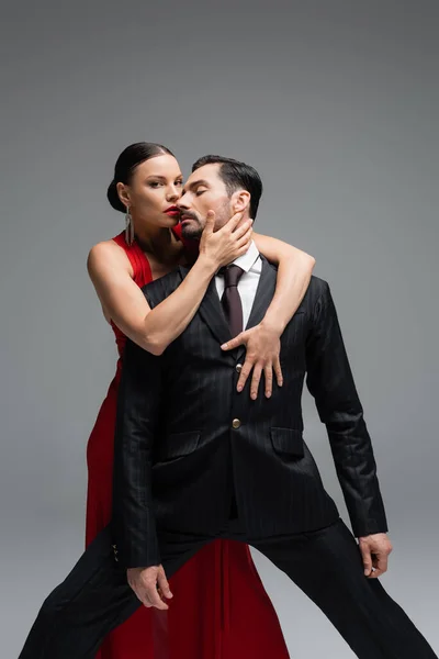 Femme élégante en robe rouge touchant l'homme en costume et regardant la caméra isolée sur gris — Photo de stock