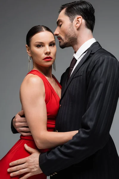 Retrato de mulher em vestido vermelho olhando para a câmera ao abraçar namorado elegante isolado no cinza — Fotografia de Stock