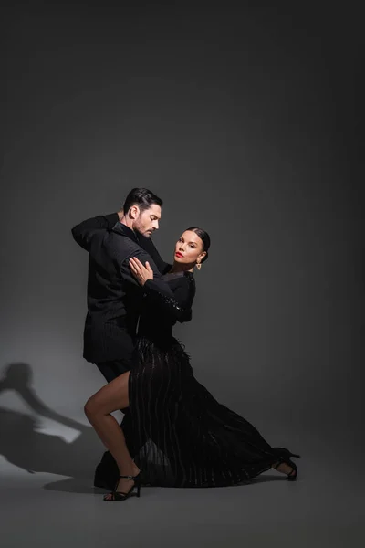Mulher elegante com lábios vermelhos olhando para a câmera enquanto dança tango com parceiro no fundo cinza com sombra — Fotografia de Stock