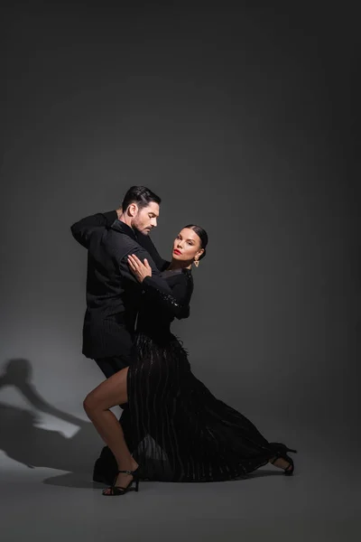 Elegant woman with red lips looking at camera while dancing tango with partner on grey background with shadow — Stock Photo