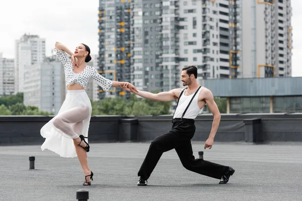 Hombre sonriente cogido de la mano de la pareja mientras baila en el techo del edificio al aire libre - foto de stock