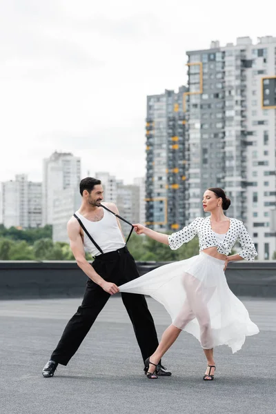 Stylish woman in heels dancing with smiling partner on roof of building outdoors — Stock Photo