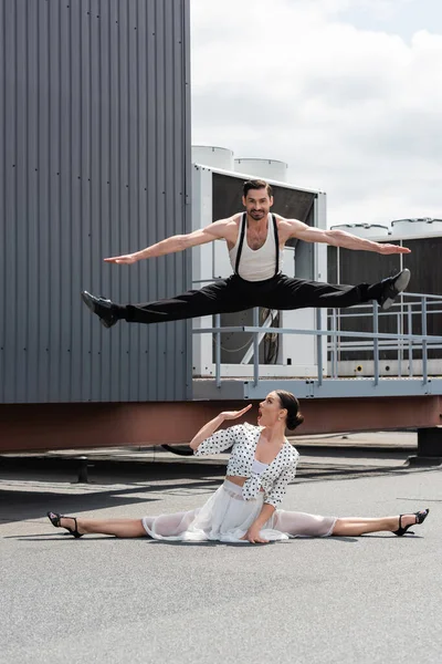 Smiling dancer doing split while jumping above shocked partner on roof of building outdoors — Stock Photo