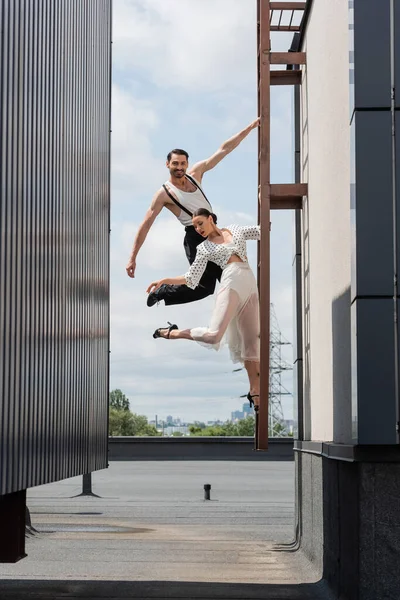 Smiling dancer looking at camera while posing near partner in skirt and heels on ladder on roof — Stock Photo