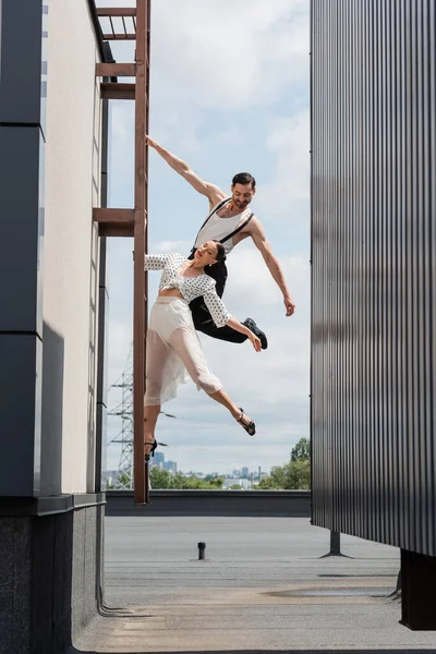 Smiling professional dancer looking at partner in heels posing on ladder on rooftop of building outdoors — Stock Photo