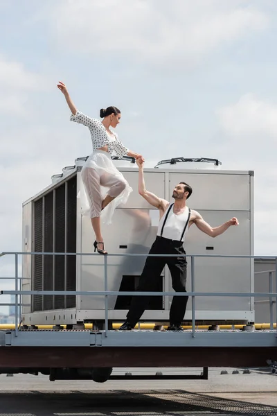 Bailarina sonriente en tacones y falda cogida de la mano del compañero en el techo del edificio al aire libre — Stock Photo